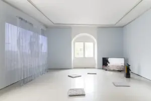 Bright room with a large left-wall screen, a TV showing the artist in the bottom right, and cushions arranged on the floor.